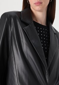 Blazer in pelle nera con colletto a revers, caratterizzato da una texture liscia e una vestibilità su misura, indossato sopra un top nero a pois con paillettes.