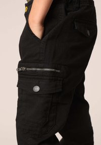 Zwarte cargobroek met meerdere zakken, een zij-rits zak en een knoopaccent. Gemaakt van stevige stof met een gladde textuur.
