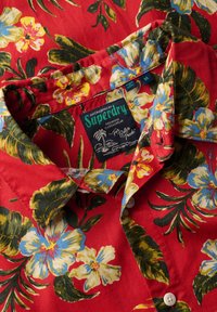 Czerwona koszula z krótkim rękawem w kwiatowy wzór, z dużymi niebieskimi i żółtymi kwiatami oraz zielonymi liśćmi, z metką "Superdry". Widoczne guziki.