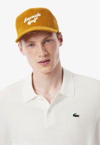 Cappello in corda senape con "Lacoste Golf" ricamato in bianco. Il modello indossa una polo bianca con il logo del coccodrillo verde sul petto.