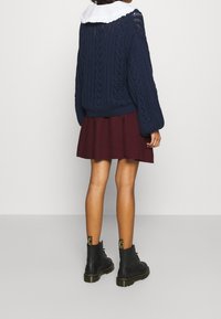 Pull en maille bleu marine avec un col à volants blanc, associé à une jupe évasée bordeaux et des bottines noires à lacets.