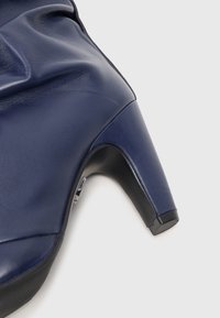 Botine cu toc înalt din piele bleumarin, cu un design curbat, având o textură netedă și o bază neagră. Accente metalice notabile aproape de toc.