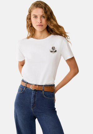 Giovane donna con capelli biondi ricci, che indossa una maglietta bianca con una spilla decorativa e jeans blu con cintura marrone, mani dietro la schiena.