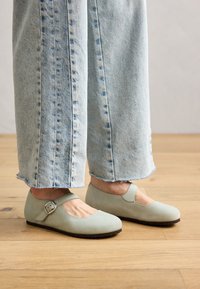Zapatos planos Mary Jane de color verde claro con punta redondeada y correa con hebilla; material de cuero suave. Combinados con jeans de pierna ancha desgastados de color azul claro.