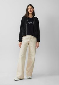 Veste bomber noire à texture côtelée, portée sur un t-shirt graphique noir. Un pantalon large beige clair et des baskets grises complètent la tenue.