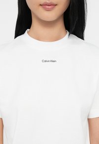 Valge puuvillane T-särk ümmarguse kaelusega, millel on rinnal must "Calvin Klein" logo, sile tekstuur ja lühikesed varrukad.