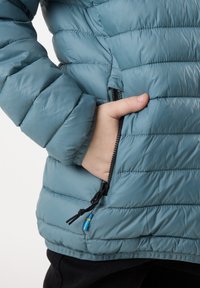 Blå vadderad jacka med horisontellt quiltat mönster, sidoficka med dragkedja och svart hårdvara, samt justerbar dragsko i nederkant.