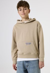 Beige Kapuzenpullover mit einer Vordertasche, versehen mit blauen Textdetails. Der Stoff hat eine weiche Textur und wird mit hellen Jeans kombiniert.