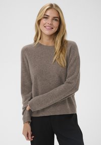 Pull marron tricoté avec encolure ronde, manches longues, poignets côtelés et texture douce, associé à un pantalon noir.