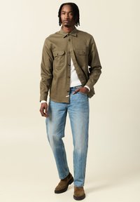 Camicia button-up verde oliva con due tasche sul petto, abbinata a jeans azzurri chiari e stivaletti alla caviglia in pelle scamosciata marrone. Design semplice e informale.