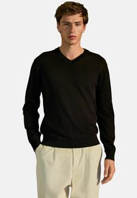 Schwarzer V-Ausschnitt-Pullover aus glattem Strickstoff, mit langen Ärmeln und geripptem Saum, kombiniert mit hellen, locker sitzenden Hosen.