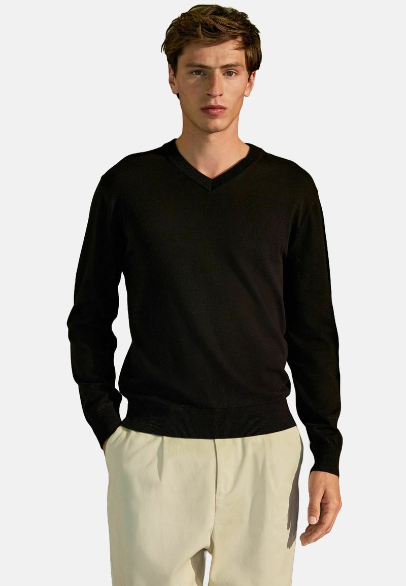 Schwarzer V-Ausschnitt-Pullover aus glattem Strickstoff, mit langen Ärmeln und geripptem Saum, kombiniert mit hellen, locker sitzenden Hosen.