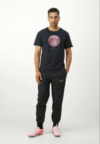 Mand står iført en sort Paris Saint-Germain Nike t-shirt, sorte Nike joggers og lyserøde Nike fodboldstøvler på hvid baggrund.