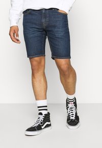 Tummansiniset farkkushortsit, joissa on viisi taskua ja näkyvä ompelu. Yhdistetty mustiin korkeisiin tennareihin ja valkoisiin raidallisiin sukkiin.