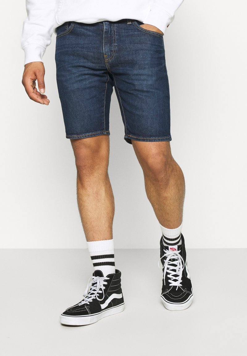 Tummansiniset farkkushortsit, joissa on viisi taskua ja näkyvä ompelu. Yhdistetty mustiin korkeisiin tennareihin ja valkoisiin raidallisiin sukkiin.