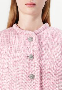 Růžový tkaný kabát s texturovanou látkou ve vzoru rybí kosti, kulatým límcem a třemi kovovými knoflíky.