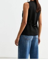 Černý bezrukávový top s detailem na zavazování vzadu, spárovaný s modrými džínovými kalhotami s vysokým pasem, které mají dvě zadní kapsy a volný střih.