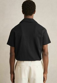 Cămașă neagră cu mâneci scurte și guler, confecționată dintr-un material neted, asortată cu pantaloni de o culoare deschisă, care au buzunare la spate și detalii cu nasturi.