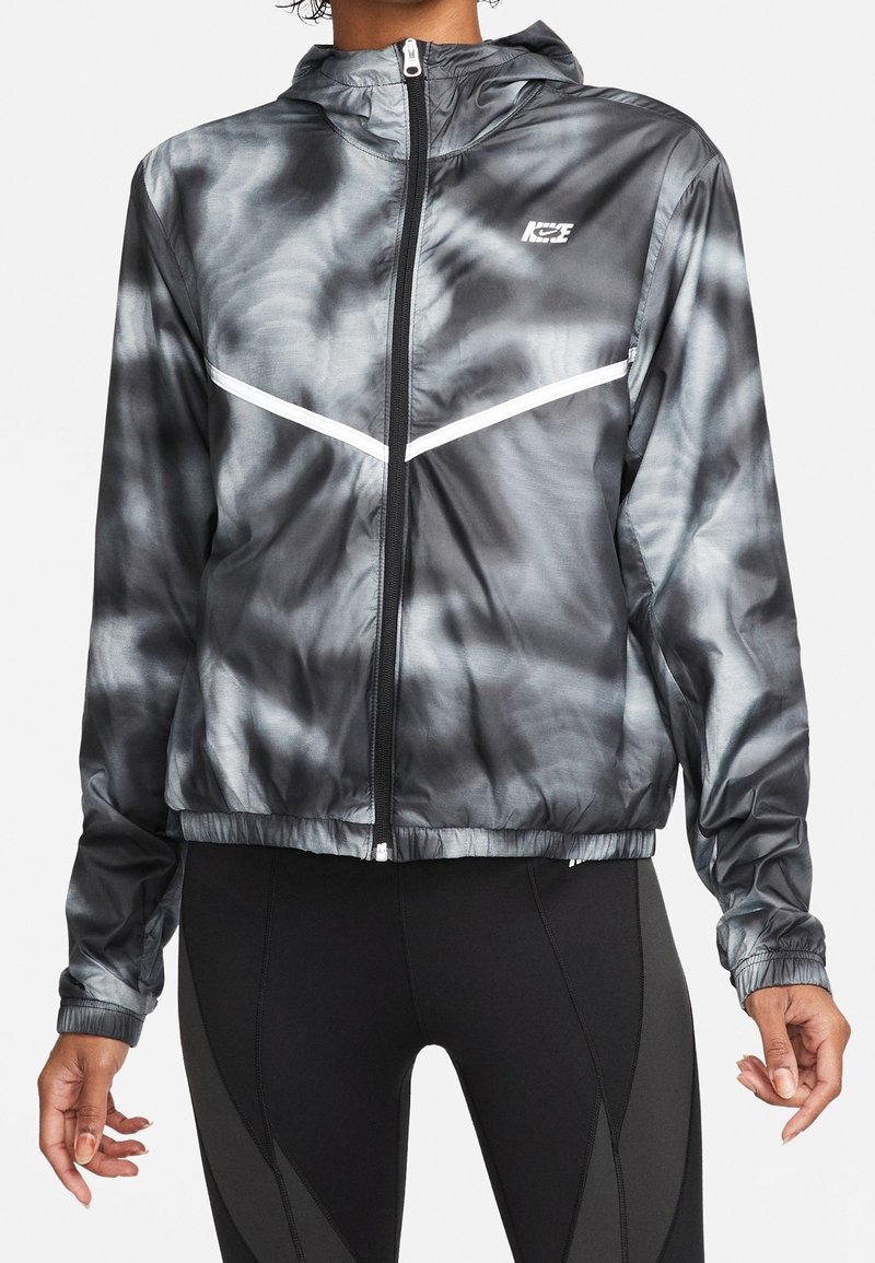 Personne portant une veste zippée Nike tie-dye noire et grise avec des bandes chevron blanches et une capuche, associée à des leggings noirs.