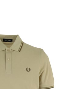 Beige poloshirt gemaakt van gestructureerde stof, met een knoopsluiting met twee knopen en korte mouwen. Bruine afwerking en geborduurd logo op de borst.