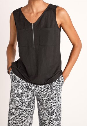 Sleeveless black top with front zipper and chest pockets paired with black-and-white patterned pants, hands in pockets, bracelet on wrist.