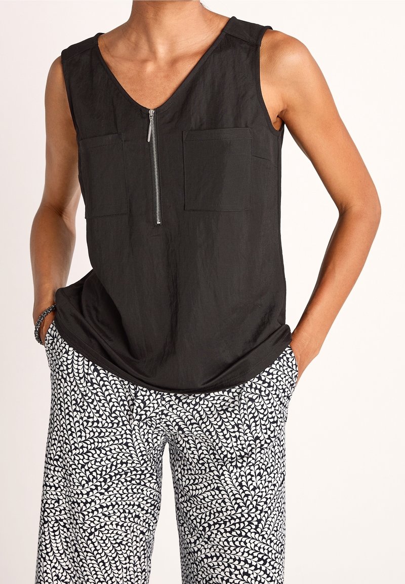 Haut noir sans manches avec fermeture éclair à l'avant et poches poitrine, associé à un pantalon à motifs noir et blanc, mains dans les poches, bracelet au poignet.