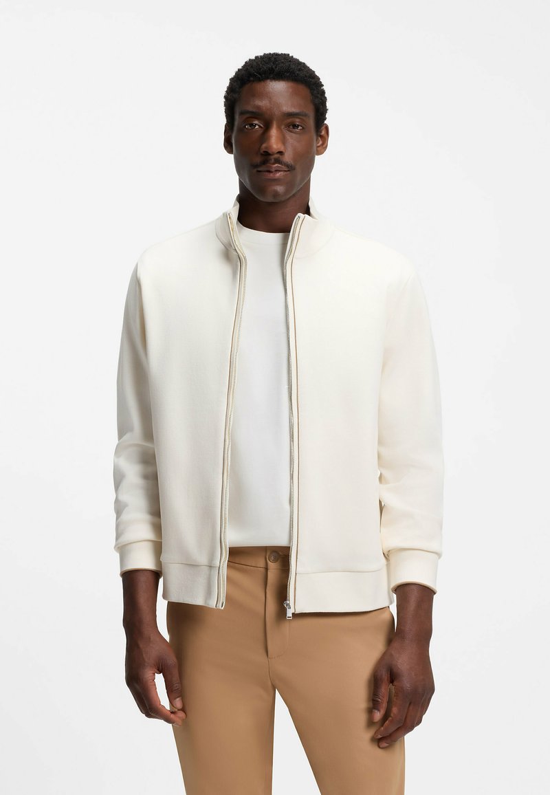 Homme portant une veste blanche à fermeture éclair sur une chemise blanche et un pantalon beige, debout devant un fond blanc uni.