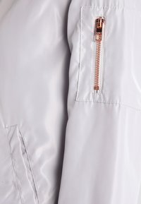 Veste bomber gris clair avec une texture lisse et brillante. Comprend une poche latérale avec une fermeture éclair en or rose et des accents cousus.