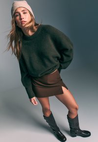 Pull oversize vert foncé, mini-jupe marron avec fente latérale et bottes marron montantes avec accents à boucle. Texture lisse dans l'ensemble.