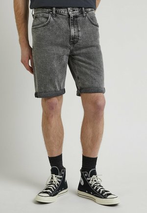 Homme portant un short en denim noir délavé retroussé, des baskets montantes noires et des chaussettes noires, debout devant un fond clair uni.
