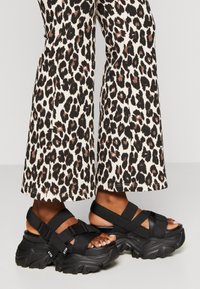 Sandales à plateforme noires avec semelles épaisses à texture détaillée, sangles réglables et design à bande croisée. Portées avec un pantalon large à imprimé animalier.