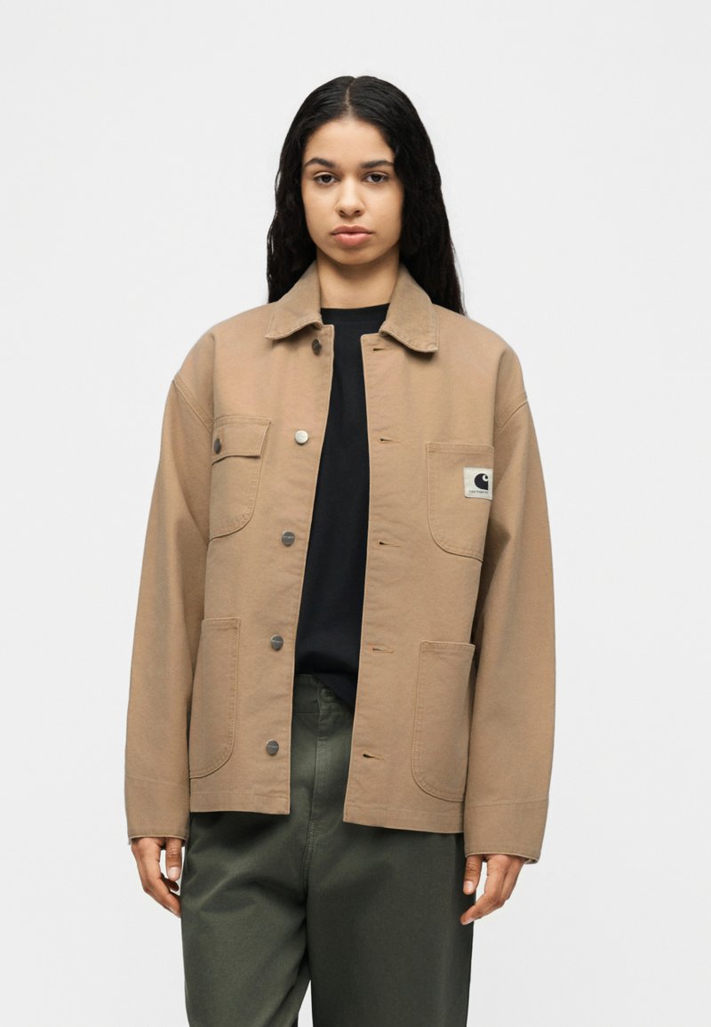 Jonge persoon met lang donker haar draagt een lichtbruine jas met knopen over een zwart shirt en olijfgroene broek, staand tegen een effen witte achtergrond.