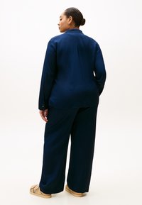 Μπλε ναυτικό μεταξένιο πουκάμισο με μακριά μανίκια και πιέτα στην πλάτη, σε συνδυασμό με παντελόνι φαρδιάς γραμμής. Το σύνολο ολοκληρώνεται με καφέ παπούτσια τύπου slip-on.