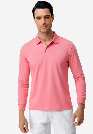 Mann, der ein pinkes langärmliges Polohemd mit Kragen und Manschetten trägt, kombiniert mit weißen Hosen, vor einem einfarbigen Hintergrund stehend.