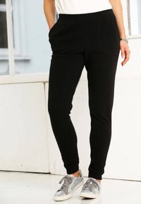 Een persoon in zwarte, strakke broek met zakken en zilverkleurige, gestructureerde sneakers staat binnen bij een witte muur en een raam.