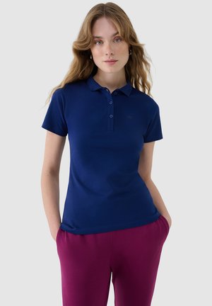 Młoda kobieta o falowanych blond włosach, ubrana w granatową koszulkę polo z krótkim rękawem i magenta spodnie, z rękami w kieszeniach.