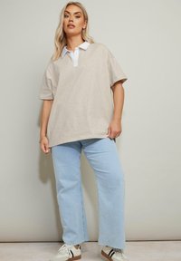 Beige oversized polo skjorta med vit krage, i kombination med ljusblå vida jeans och vita sneakers med svarta och bruna accenter.