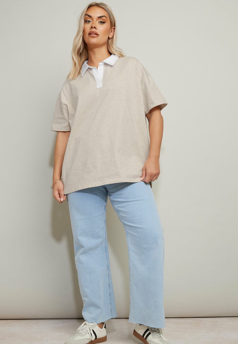 Beige oversized polo skjorta med vit krage, i kombination med ljusblå vida jeans och vita sneakers med svarta och bruna accenter.