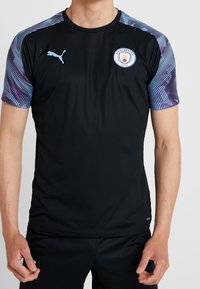 Musta lyhythihainen urheilupaita, jossa on siniset ja violetit raidat olkapäillä, varustettuna kudotulla Manchester City -logolla rinnassa.