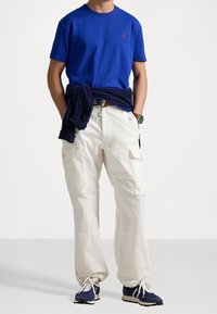 Camiseta de algodón azul con un pequeño logo, pantalones cargo blancos con bolsillos laterales y zapatos deportivos azules con suela blanca y detalles en blanco.