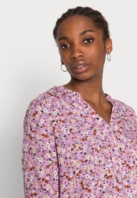 Blusa floreale in viola chiaro con una consistenza morbida, caratterizzata da scollatura a V, chiusura a bottoni e un motivo di piccoli fiori multicolore.