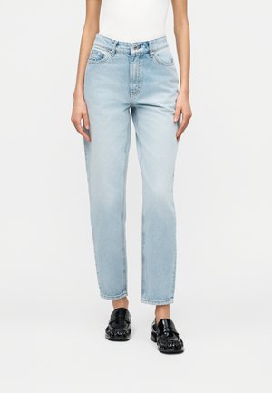 Persoon draagt lichtblauwe rechte jeans met hoge taille, wit mouwloos topje en zwarte glanzende loafers, staand tegen een effen witte achtergrond.