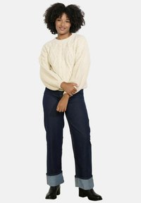 Krémový sveter s vrkočovým vzorom a nadýchanými rukávmi, skombinovaný s tmavomodrými džínsami s vyhrnutými lemami a čiernymi členkovými čižmami.