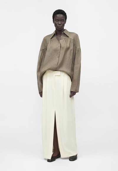 Γυναίκα που φοράει μια χαλαρή καφέ μπλούζα με κουμπιά και μακριά λευκή φούστα με σκίσιμο μπροστά, στέκεται μπροστά σε ένα απλό λευκό φόντο.