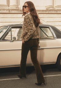 Leopardenmuster Kunstpelzjacke, braune Corduroy Schlaghosen, schwarze High Heels und dunkle Sonnenbrillen vor dem Hintergrund eines Oldtimers.