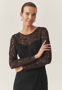 Rochie neagră din dantelă, cu modele florale complicate, mâneci lungi și decolteu rotund. Textura pare delicată și stratificată. Cercei aurii în formă de cerc.
