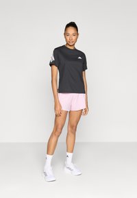 Camiseta deportiva negra con logo blanco, combinada con pantalones cortos rosas. El modelo lleva zapatos blancos con detalles en rosa y calcetines blancos. Fondo neutro.