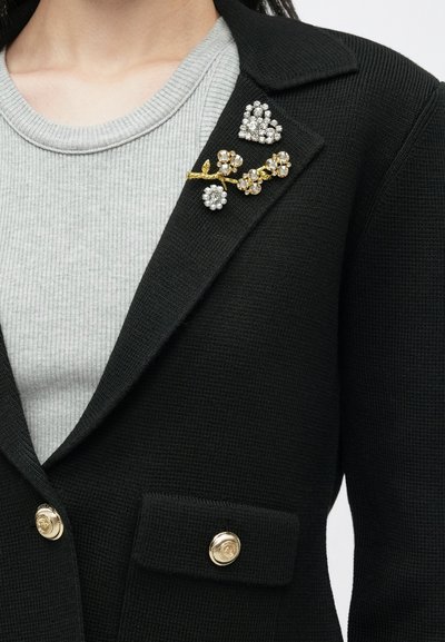 Μαύρο σακάκι με υφή και χρυσά μπουτόν, που διαθέτει μια διακοσμητική καρφίτσα με λαμπερά ασημί και χρυσά λουλουδάτα στοιχεία σε έναν γιακά.