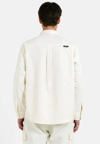 Camisa de manga larga en tejido texturizado color blanco roto con un pliegue en la parte trasera. Presenta un cuello puntiagudo y una pequeña etiqueta con el logo cerca del escote.