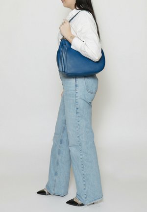 Donna con camicia bianca e jeans azzurri chiari che porta una borsa a tracolla in pelle blu con nappe su una spalla, su sfondo semplice.